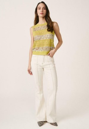 Mujer con top de punto sin mangas amarillo con rayas metálicas, combinado con pantalones blancos acampanados de talle alto y zapatos puntiagudos.