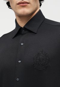 Άνδρας που φοράει μαύρο πουκάμισο με κουμπιά και μυτερό γιακά, με ένα διακριτικό κεντητό έμβλημα στο αριστερό στήθος.