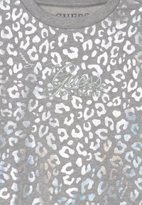 T-shirt grigio a maniche corte con un motivo maculato argento e un logo "Guess" riflettente sulla parte anteriore. Materiale in cotone morbido.