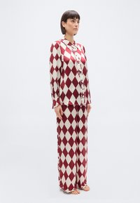 Femme debout, pieds nus, portant un chemisier à manches longues à motifs losanges rouges et blancs ainsi qu'un pantalon large assorti, sur un fond uni.