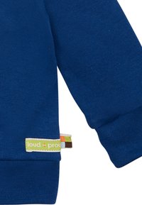 Felpa blu con una texture liscia, polsini a costine e un'etichetta che presenta "loud + proud" in verde. L'etichetta ha strisce multicolori.