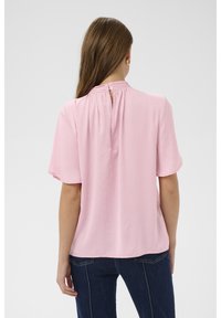 Blusa rosa a maniche corte con un'apertura a goccia sulla schiena, che presenta un motivo a puntini in rilievo e un design fluente. Indossata con jeans blu.
