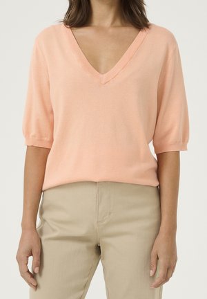Femme portant un pull léger à col en V et manches courtes couleur pêche clair, et un pantalon beige, debout avec les bras détendus le long du corps.