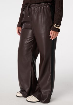 Bruine nepleder wijde broek met een trekkoord in de taille, voorzien van zwarte zijstrepen. Gecombineerd met witte instap schoenen met studs.