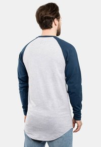 Pitkähihainen paita, jossa on harmaa runko ja tummansiniset raglan-hihat. Sileä kangas, pyöristetty helma ja hienovarainen logotyyli takana kaula-aukossa.