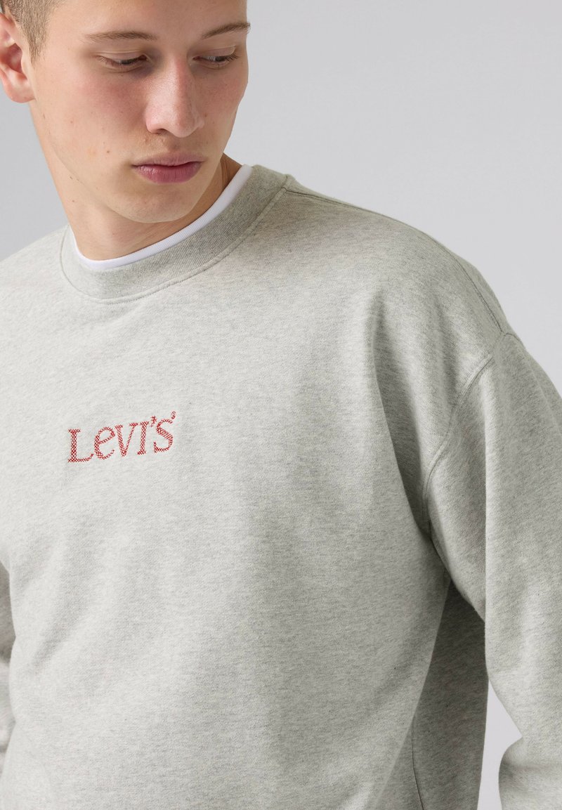 Giovane uomo che indossa una felpa Levi's grigio chiaro con ricamo del logo rosso, guarda verso il basso e leggermente di lato su uno sfondo semplice.