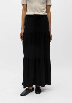 Kobieta ubrana w beżową bluzkę z krótkim rękawem i długą, czarną, warstwową spódnicę ze ściągaczem w pasie, w parze z czarnymi wsuwanymi butami.