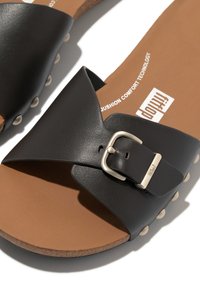 Zwarte leren slippers met een brede band voorzien van een zilveren gesp, een zachte bruine binnenzool en lichtgekleurde accentstuds langs de randen van de zool.