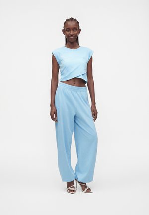 Femme debout portant un haut court sans manches bleu clair, un pantalon ample assorti et des sandales blanches à talons hauts sur un fond uni.