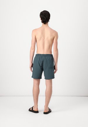 Młody mężczyzna stoi boso, odwrócony plecami, ubrany w ciemnoniebieskie kąpielówki i czarne sandały, na jednolitym jasnym tle.