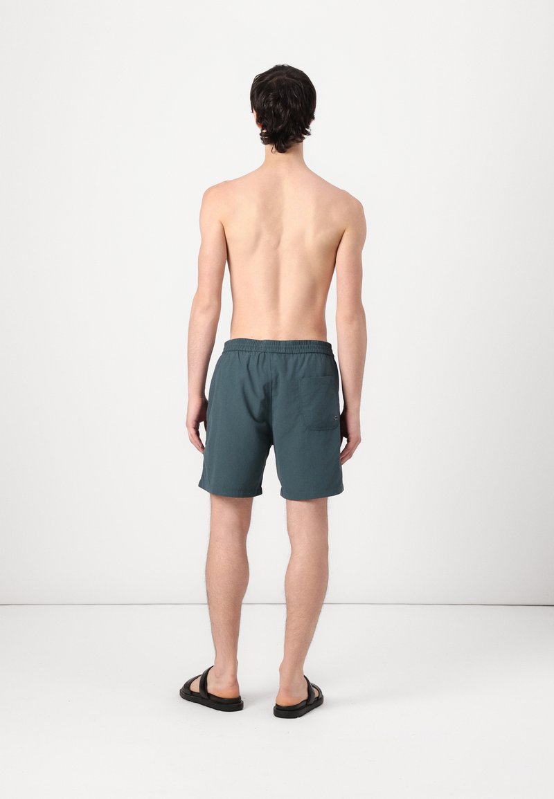 Jeune homme debout pieds nus, tourné vers l'arrière, portant un short de bain bleu sarcelle foncé et des sandales noires, sur un fond clair uni.