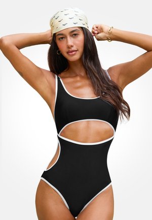 Vrouw in een zwart éénstukbadpak met witte bies en uitsnijdingen bij de taille, die een crèmekleurige hoofddoek met kleine zwarte patronen rechtzet.