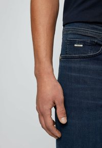 Jeans en denim bleu foncé avec une coupe slim. Les caractéristiques incluent une étiquette de marque sur la poche arrière et une texture lisse. Main reposant de manière décontractée.