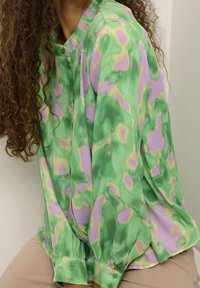 Mujer con cabello rizado que lleva una blusa de patrón abstracto en verde, lavanda y beige con botones y pantalones beige, sentada contra un fondo liso.