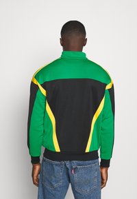 Grön och svart jacka med gula detaljer, hög krage, elastiska ärmslut och sidopaneler som visar en sportig design och en texturerad tyg.