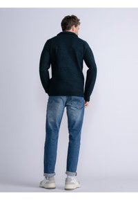 Maglione blu scuro con colletto, polsini e orlo a coste. Abbinato a jeans blu chiaro e scarpe da ginnastica bianche, visto da dietro.