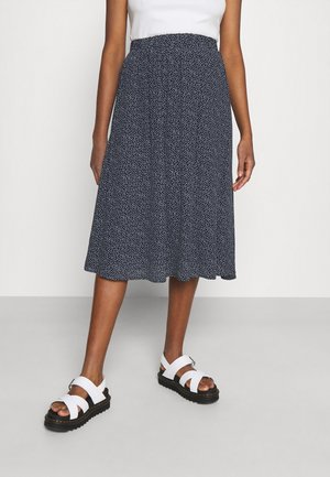 Femme portant une jupe midi bleu marine à pois blancs, un haut sans manches blanc et des sandales blanches à semelles noires, debout sur un sol gris.