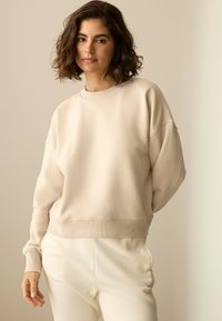 Bež dukserica s rebrastim okruglim izrezom i spuštenim ramenima, uparena s bijelim trenirkama koje imaju diskretan logo sa strane.