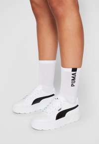 Sneakers atletici bianchi con accenti neri, suola in gomma testurizzata e colletto imbottito, abbinati a calzini bianchi con il logo "PUMA" nero.
