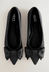 Ballerines en satin noir avec un bout pointu et un accent en nœud à l'avant. Texture lisse avec une finition brillante sur le matériau.