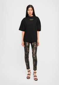 Young woman wearing black oversized "Just Cavalli True Lover" T-shirt, snake print leggings, and black strappy high-heeled sandals against a white background.