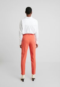 Úzké korálové kalhoty s vysokým pasem, v kombinaci s bílou pruhovanou košilí. Oblečení má minimalistický design a čisté linie.