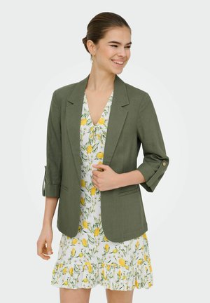 Uśmiechnięta kobieta z włosami w kok nosi oliwkową marynarkę na białej sukience z żółtymi cytrynami i zielonymi liśćmi, stojąc na jednolitym tle.