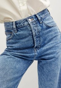 Jeans de mezclilla azul de tiro alto con un acabado descolorido, cierre de botón en la parte delantera y cinco bolsillos, que presentan detalles clásicos de costura.