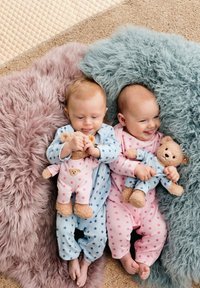Two infants lie on textured faux fur blankets, wearing pastel-colored onesies with star patterns. Each holds a plush bear toy.