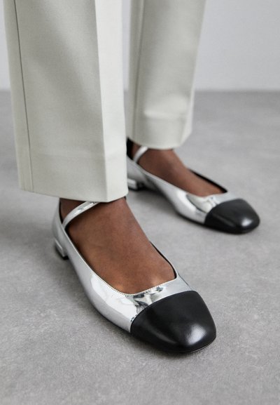Ballerine metallizzate argento con punta in pelle nera. Suola piatta e design con cinturino, posizionate accanto a pantaloni chiari su una superficie grigia.