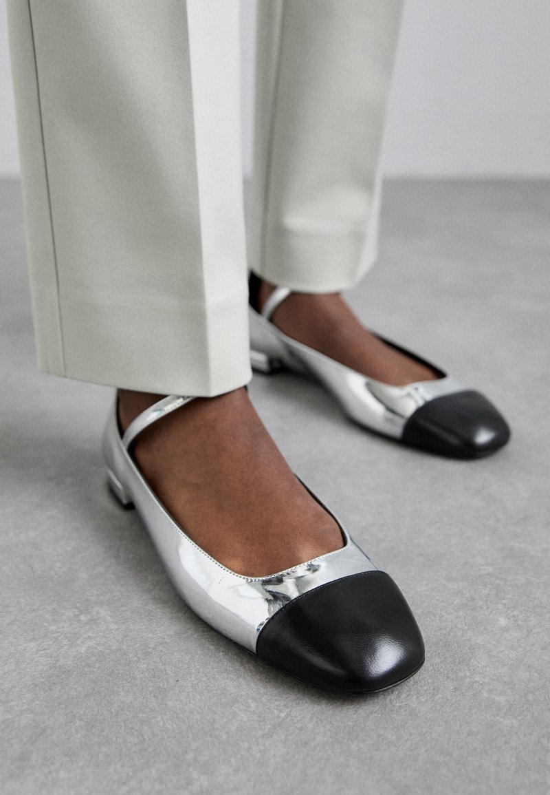 Silberne metallische Ballerinas mit einer Kappe aus schwarzem Leder. Flache Sohle und Designeriemen, platziert neben hellen Hosen auf einer grauen Oberfläche.