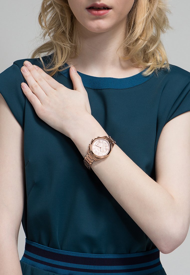 Reloj de pulsera color oro rosado con una cara redonda, que presenta una esfera clara y múltiples marcas. El reloj está en una muñeca con un tono de piel suave y claro.