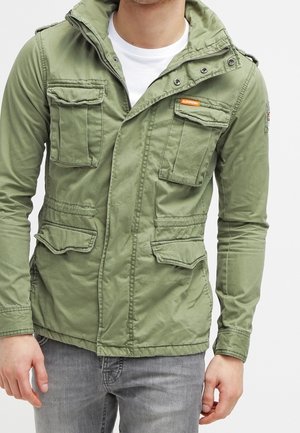 Mężczyzna ubrany w jasnooliwkową kurtkę użytkową z czterema przednimi kieszeniami na klapki, zamkiem błyskawicznym i zapięciem na zatrzaski, pod którą ma białą koszulkę.