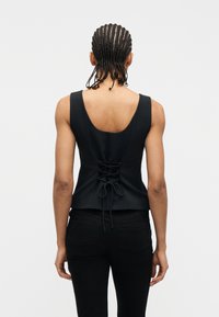 Άτομο με πλεξούδες που φοράει μαύρο αμάνικο τοπ με κορδόνια στο πίσω μέρος και μαύρο παντελόνι, στέκεται μπροστά σε απλό φόντο.