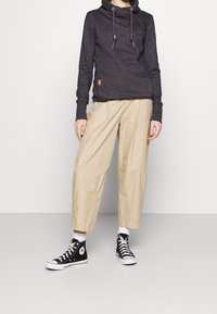 Felpa con cappuccio grigio scuro con coulisse, abbinata a pantaloni beige larghi e sneakers nere alte. Tessuto morbido, vestibilità casual.