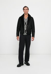 Hombre con chaqueta de shearling negra, camisa de patrón blanco y negro, pantalones cargo negros y zapatos de cuero negros de pie contra una pared blanca.