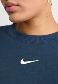 Sweatshirt azul marinho com decote canelado, apresentando um logotipo branco da Nike na frente. A pele exibe uma pequena tatuagem de coração vermelho no pescoço.