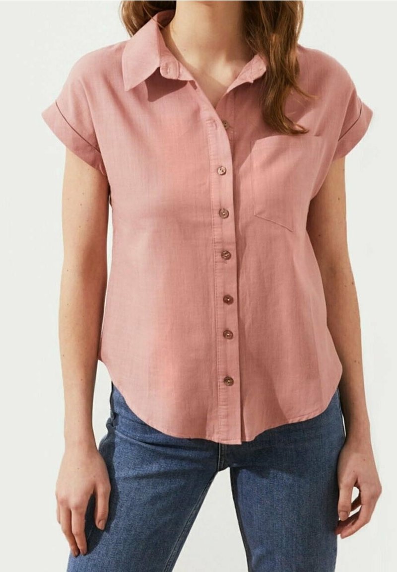Chemise rose clair à manches courtes retournées, avec poche avant et ourlet arrondi. Le tissu semble lisse avec une texture subtile.