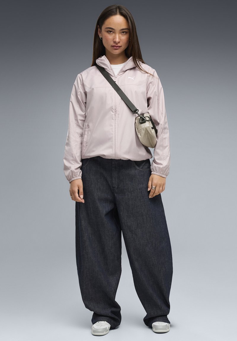 Femme portant une veste zippée rose clair, un jean large foncé, des baskets blanches et un petit sac bandoulière beige sur un fond gris.
