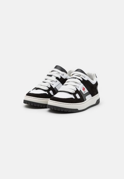 Sneaker in bianco e nero con esterno in suede e pelle, lacci bianchi piatti, suola in gomma e un piccolo dettaglio con logo rosso.