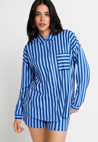 Pijama set u plavim i bijelim prugama, koji uključuje košulju na dugmad s džepom na grudima i odgovarajuće kratke hlače. Pamuk, opušteni kroj.
