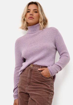 Lys lilla turtleneck sweater med ribbet tekstur, lange ærmer og afslappet pasform, båret med brune fløjlsbukser med en frontlomme.