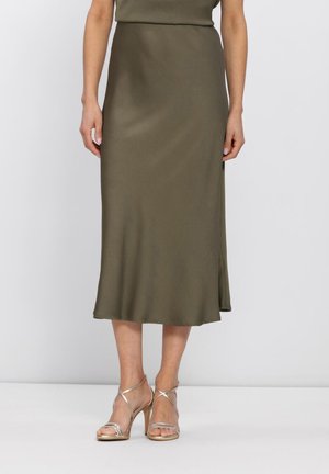 Frau trägt einen olivgrünen Satin-Midirock, kombiniert mit goldfarbenen, riemchenbesetzten High-Heel-Sandalen, stehend vor einem schlichten Hintergrund.