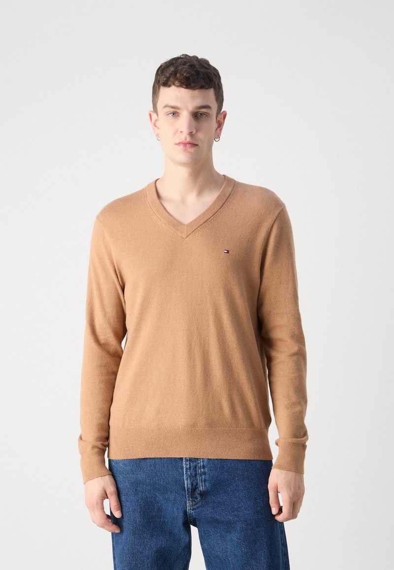 Maglione marrone a scollo a V realizzato in materiale morbido, con maniche lunghe e orlo a coste. Dettaglio del logo piccolo sul lato sinistro del petto. Indossato sopra jeans blu.