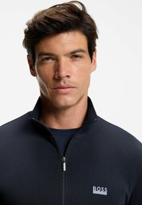 Musta vetoketjullinen takki, jossa korkea kaulus ja hillitty logo. Sileä koostumus, istuva malli ja näkyvissä oleva kontrastivärinen tummansininen aluspaita.