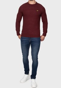 Bordeauxfarbener Strickpullover mit strukturiertem Muster, Rundhalsausschnitt und kleinem gelbem Logo. Kombiniert mit blauen Jeans und weißen Sneakern.