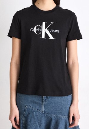 Femme portant un t-shirt noir Calvin Klein Jeans avec un grand logo « CK » blanc et gris, associé à une jupe en jean bleue.