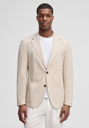 Mann trägt ein beiges Zweiknopf-Sakko über einem weißen Rundhalsausschnittshirt und weißen Hosen, steht vor einem hellgrauen Hintergrund.