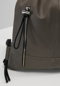 Sac à dos en cuir gris avec une finition texturée, doté d'une fermeture éclair en argent, d'un détail à cordon noir et d'une poche latérale pour le rangement.