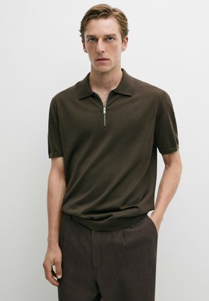 Giovane uomo con capelli chiari che indossa una polo marrone scuro a maniche corte con zip e pantaloni scuri, in piedi davanti a uno sfondo chiaro uniforme.